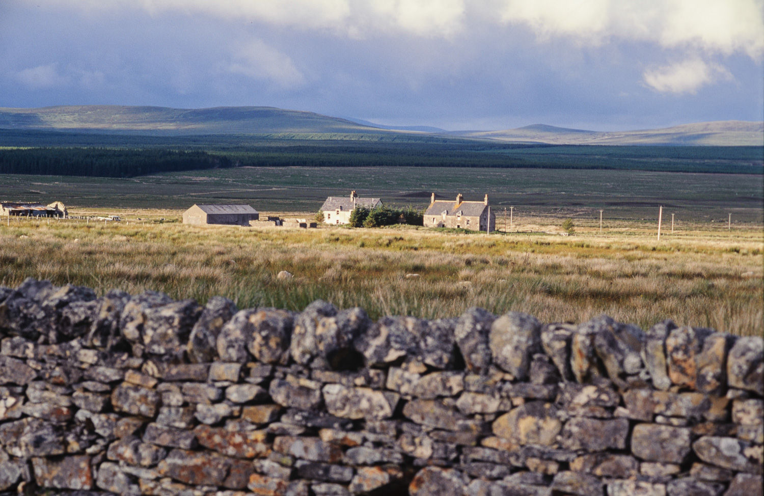 Schots landschap, een stenen muur en huisjes in de verte bij laagstaande zon.